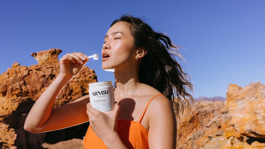 A woman outdoors in bright sunlight tipping an ARMRA Colostrum travel stick into her mouth
